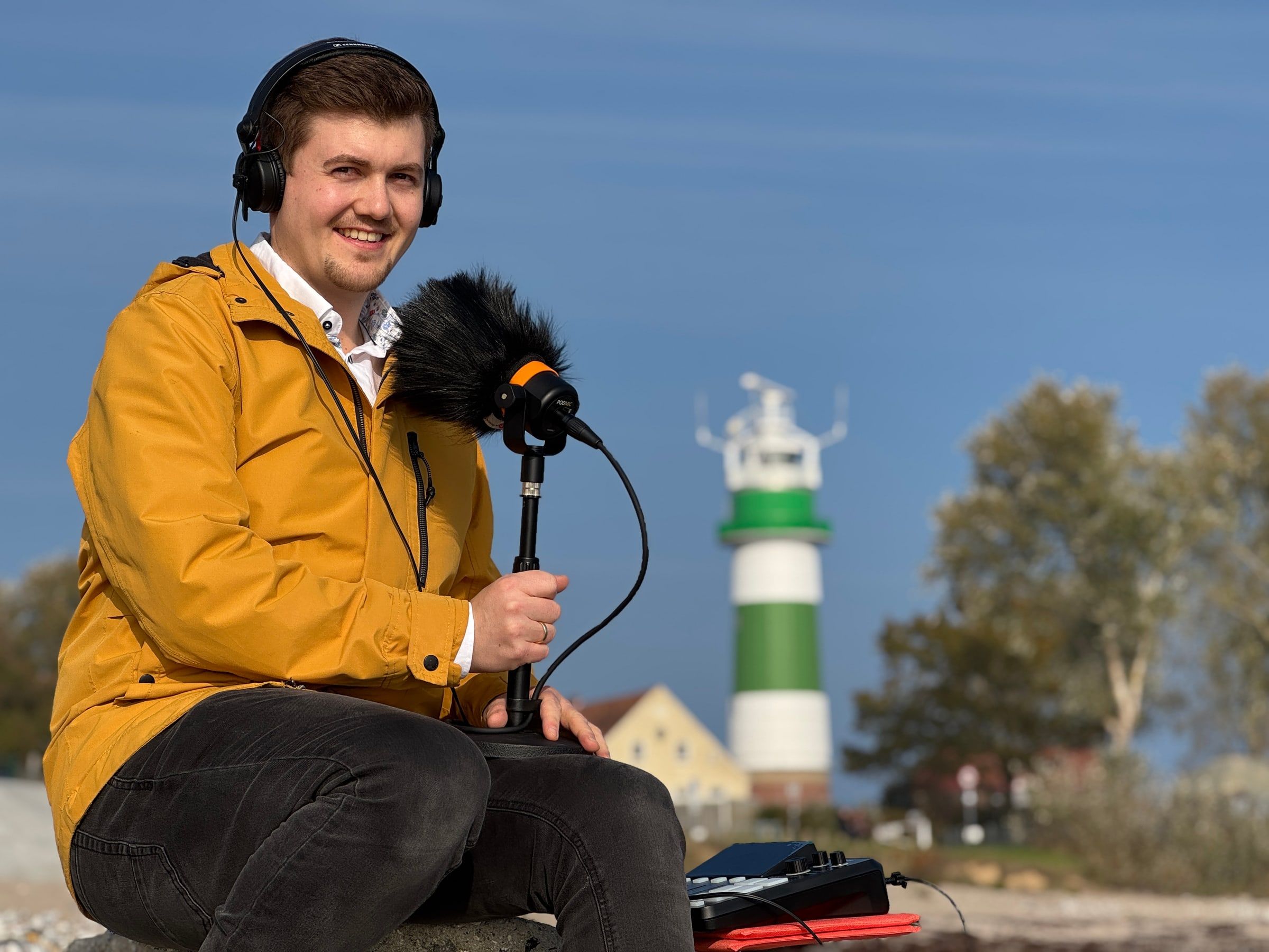 Justus sitzt mit Kopfhörern und Mikrofon am Strand, im Hintergrund ein grün-weißer Leuchtturm